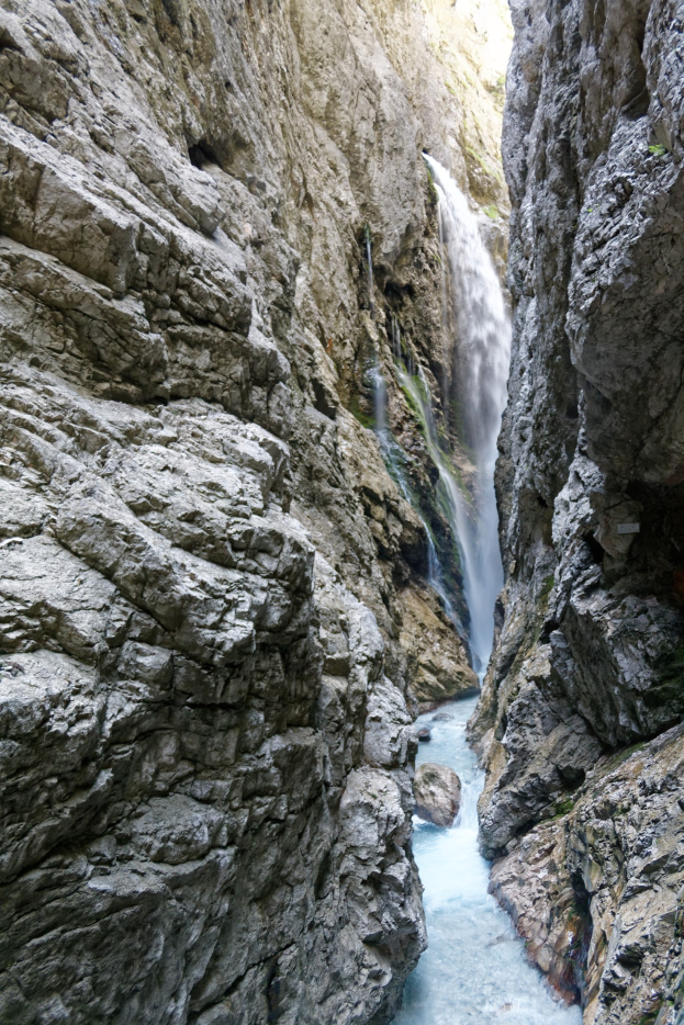 Ein kleiner Wasserfall stürzt sich über zerklüftete Felsen in einem steinigen Tal hinab, umgeben von saftig grünen Hügeln, mit klarem Wasser, das von hellem Sonnenlicht beleuchtet wird.