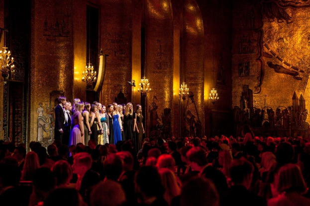 Eine Gruppe von Menschen auf einer hell erleuchteten Bühne im Royal Opera House in London mit bemalten Kulissen, die für ein Publikum unten auftreten.