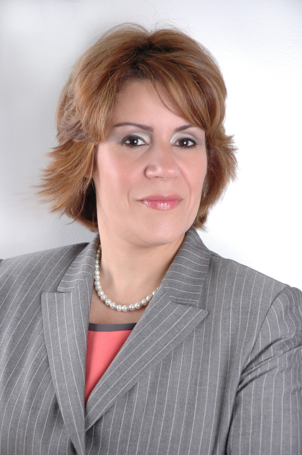 Eine professionelle Frau in einem marineblauen Anzug mit weißem Hemd und gemustertem Schlips posiert selbstbewusst vor einem weißen Hintergrund, ihre Haltung aufrecht und ihr Blick direkt.