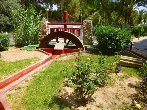 Minigolfanlage in einem Park mit Grünflächen, einer Straße mit Fußgängern und Schildern sowie einem klaren Himmel im Hintergrund.