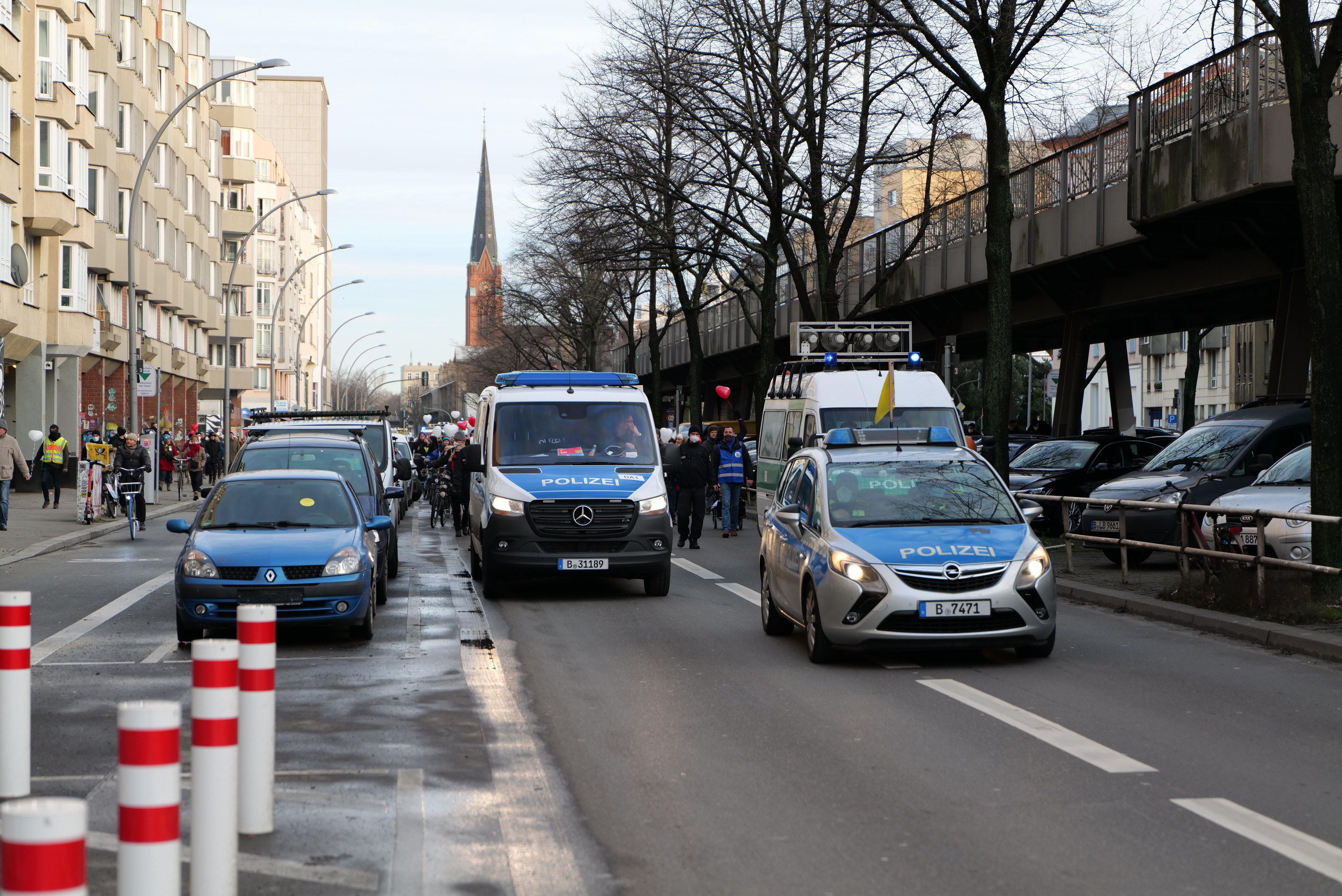 Polizeifahrzeuge fahren auf einer Straße mit hohen Gebäuden, Passanten auf dem Gehweg, Radfahrern, einer Brücke mit Geländern, Bäumen und einem klaren blauen Himmel.