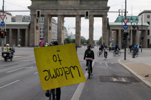 Eine Gruppe von Radfahrern mit Helmen fährt eine Straße vor dem Brandenburger Tor in Berlin, Deutschland, entlang, wobei eine Person ein gelbes Schild hält, Lichtmasten, Verkehrszeichen, Gebäude, Bäume und ein klarer blauer Himmel im Hintergrund zu sehen sind.