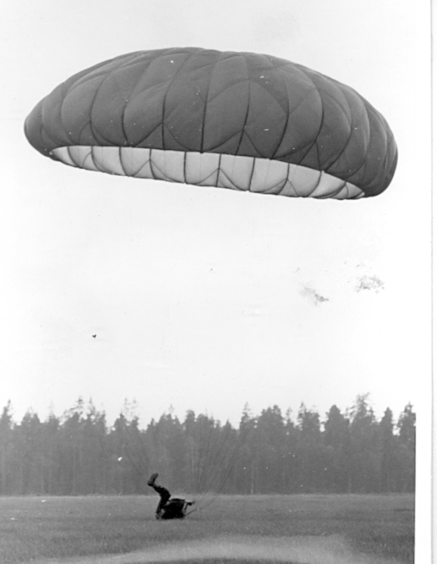 Schwarzes und weißes Foto einer Person, die mit einem Fallschirm paragleitet, umgeben von Bäumen und einem klaren Himmel, mit Text unten.