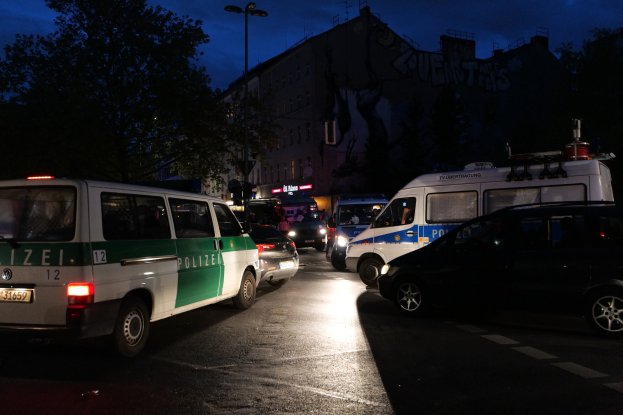 Eine Gruppe von Polizeiwagen, darunter ein Van, ein Auto und ein Rad, parkt nachts am Straßenrand mit Gebäuden, Bäumen, Laternenmasten und einem sternenklaren Himmel im Hintergrund.