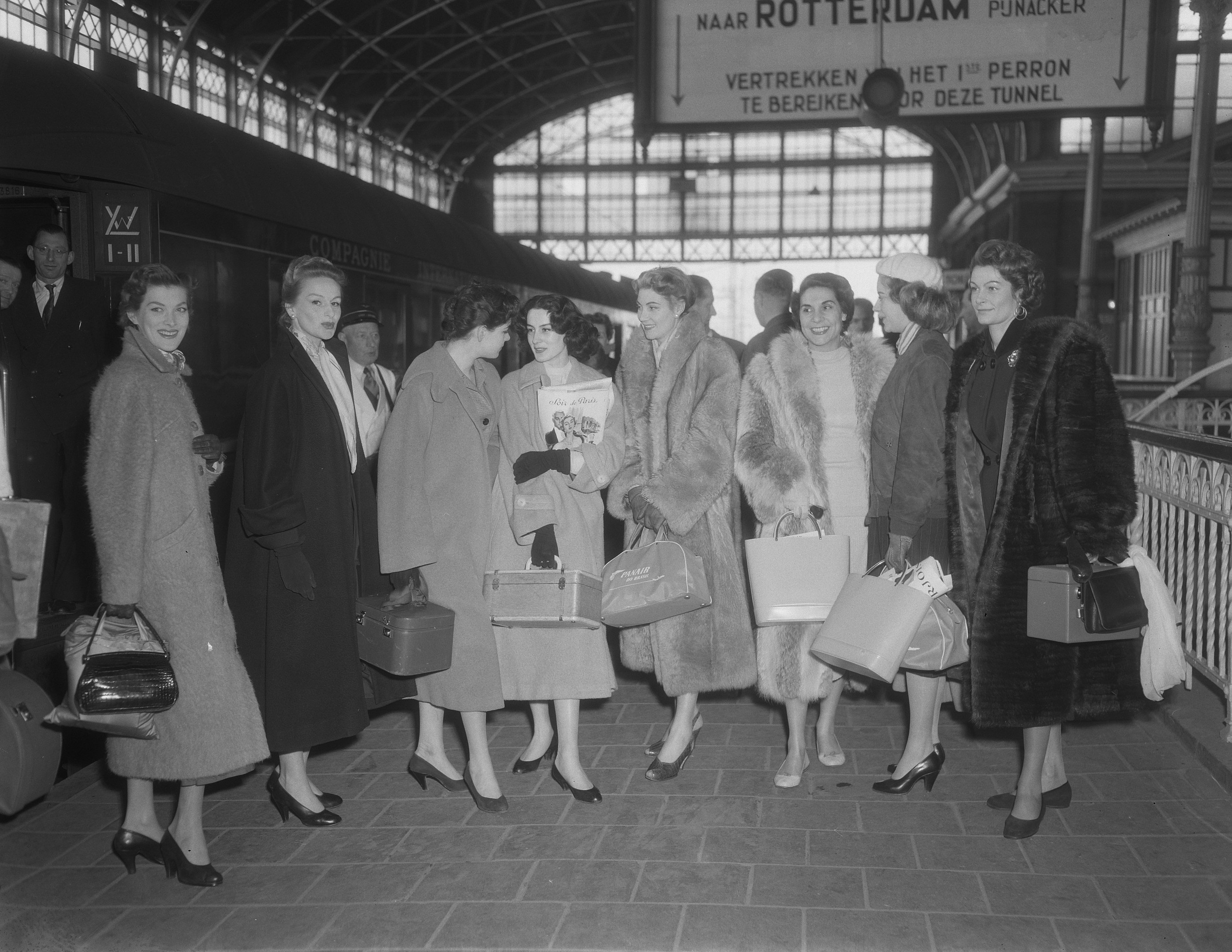 Schwarz-weiß-Bild von Frauen, die an einem Bahnhof stehen, jede hält eine Tasche, mit einem Zug auf der linken Seite und Geländern und einer Tafel auf der rechten Seite.