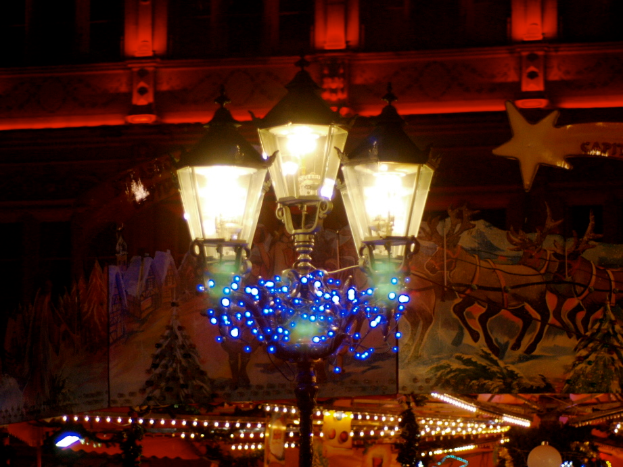 Eine Straßenlaterne in der Mitte einer Stadt bei Nacht, geschmückt mit Lichtern und Deko-Artikeln, umgeben von Gebäuden im Hintergrund, die eine festliche Atmosphäre schaffen.