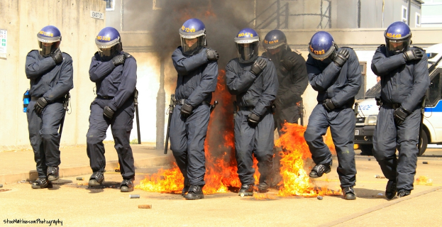 Menschen mit Helmen stehen vor einem Feuer mit verschiedenen Gegenständen auf dem Boden, Gebäude im Hintergrund, ein Fahrzeug und ein Plakat und eine Tafel an der linken Wand, mit Text am unteren Bildrand.
