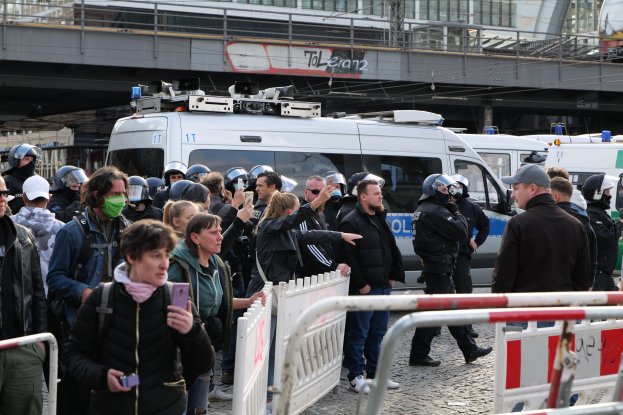 Eine Gruppe von Menschen steht vor Polizeifahrzeugen, einige tragen Helme und halten Telefone, mit Barrikaden im Vordergrund und einer Brücke und Gebäuden im Hintergrund während einer Protestauflösung in Berlin, Deutschland.