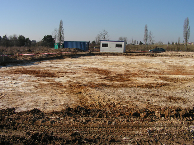 Ein schmutziger Platz mit einem Auto in der Mitte, umgeben von Bäumen, Hütten, Pfählen und anderen Gegenständen, unter einem sichtbaren Himmel, mit schlammigem Boden.
