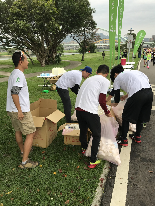 Gruppe von Menschen an einer Straße, die an einem Spendenlauf teilnehmen, umgeben von verstreuten Kartons, Flaschen und anderem Müll, mit Bäumen, Gebäuden und Schildern im Hintergrund bei klarem blauem Himmel.