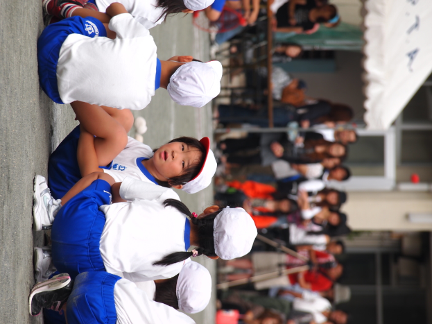 Kinder sitzen im Vordergrund auf der Straße, mit Menschen, einem Stativ, einem Zelt und einer Glaswand im Hintergrund.