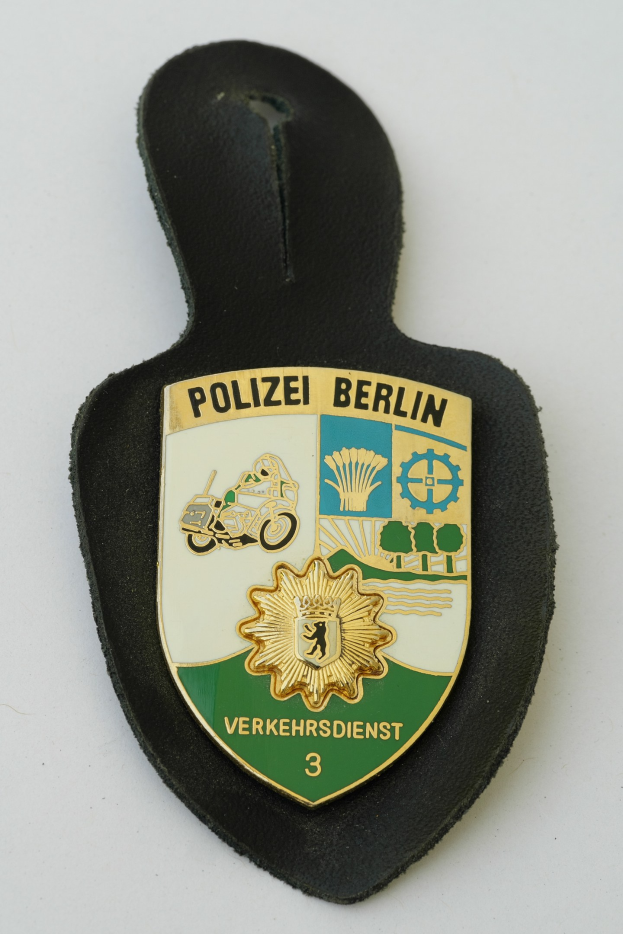 Ein schildförmiges Polizeibadged mit der Aufschrift 'Polizei Berlin' auf blauem Hintergrund, weißem Rand, zentralem weißem Stern und schwarzer fetter Schrift, das auf einer Oberfläche liegt.