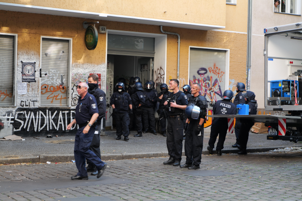 Eine Gruppe von Polizisten, einige mit Helmen und Waffen, stehen und gehen vor einem mit Graffiti bedeckten Gebäude, mit einem Fahrzeug und verschiedenen Gegenständen im Hintergrund.