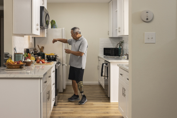 Ein Mann steht in einer Küche mit wei├čen Schr├Ąnken, h├Ąlt ein Glas in der Hand, w├Ąhrend verschiedene Gegenst├Ąnde, darunter Obst, Gemüse, Sch├Â├če und L├Âffel, auf einer Arbeitsplatte zu seiner Linken sind.