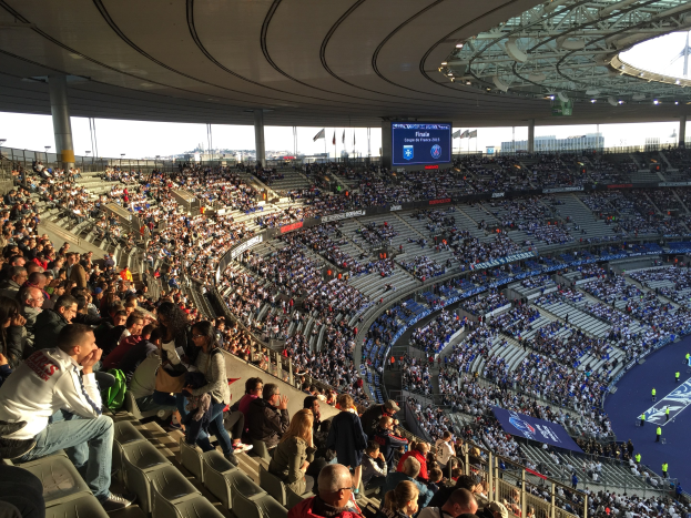Große Zuschauermenge in einem Stadion bei einem Füßtballspiel, mit einer Bühne auf der rechten Seite, Fahnen, Masten, einem Bildschirm und der Allianz Arena in München im Hintergrund.