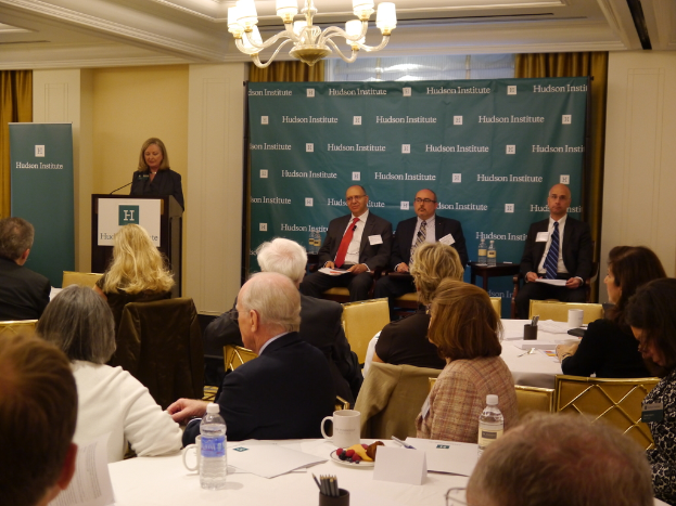 Eine Gruppe von Menschen sitzt an einem Tisch mit Wasserflaschen, Tassen und Papieren, während eine Frau an einem Rednerpult spricht.