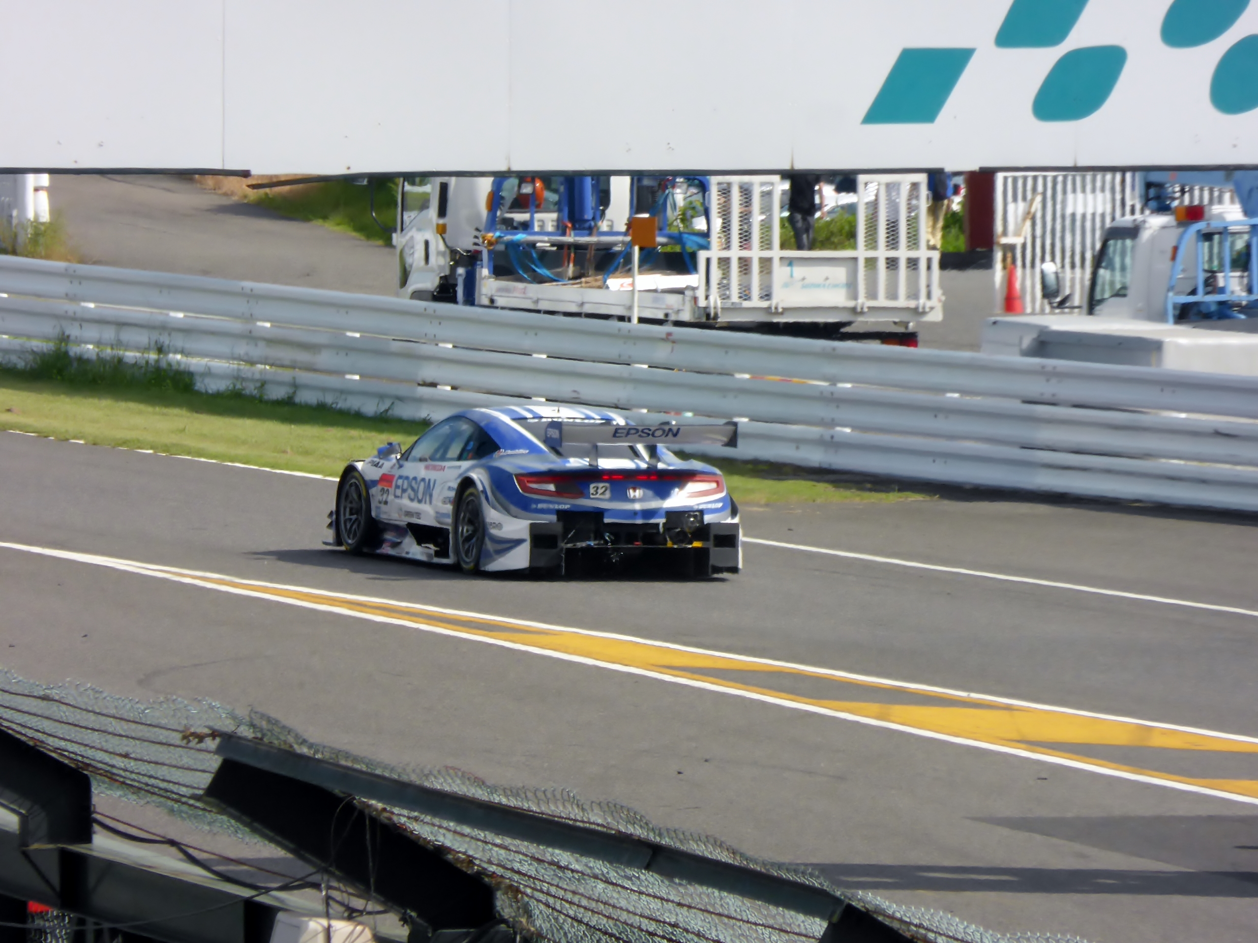 Ein BMW M6 GT3 fährt auf einer Rennstrecke bei den 24 Stunden von Le Mans, umgeben von einer Absperrung, Gras, Verkehrskegeln, Menschen und anderen Gegenständen, mit Bäumen und einem klaren blauen Himmel im Hintergrund.