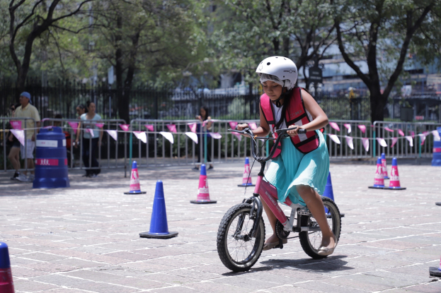 Ein junges Mädchen mit Helm und Tasche fährt auf einem Fahrrad durch eine Parkplatzmarkierung, mit einigen Menschen und Bäumen im Hintergrund.