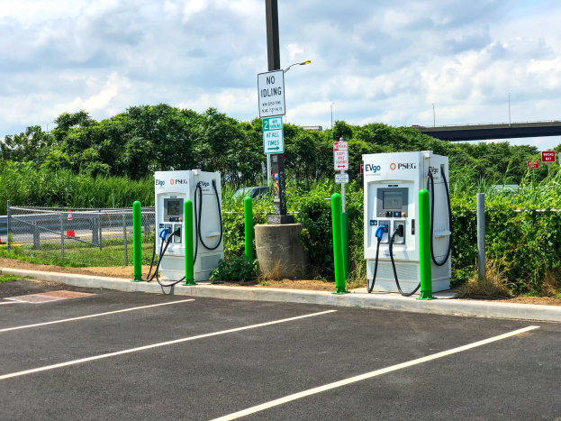 Elektrofahrzeug-Ladestation auf einem Parkplatz mit umgebender Infrastruktur, Vegetation und einer Brücke im Hintergrund unter einem bewölktem Himmel.