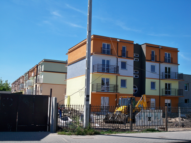 Eine Reihe von Wohngebäuden mit Fenstern, Geländern und Balkonen, einem Metallzaun, einem Pfahl, einem Bulldozer, einigen Pflanzen, einer Gruppe von Bäumen und einem bewölkten Himmel.