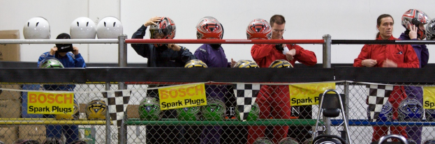 Ein Maschendrahtzaun mit Plakaten und Flaggen daran, mit ein paar Menschen im Hintergrund, einige tragen Helme, und zusätzliche Helme auf einem Ständer, alles vor einer Wand.