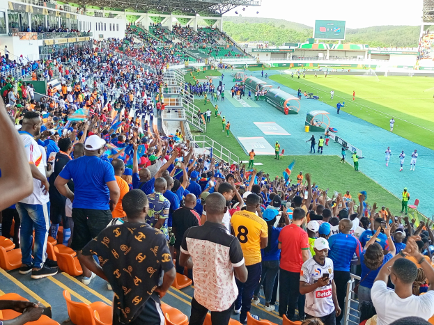 Eine große Menschenmenge steht und sitzt vor einem eingezäunten Fußballfeld mit Hügeln und einem klaren blauen Himmel im Hintergrund und wartet auf ein Spiel.