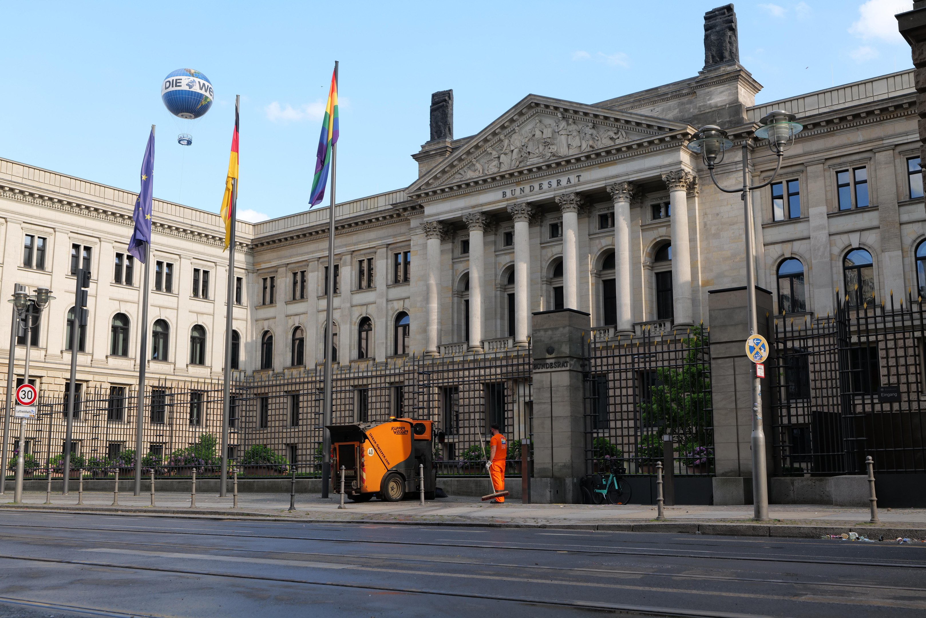 Großes modernes Gebäude mit vielen Fenstern, als Bundestag in Berlin identifiziert, umgeben von Straßeninfrastruktur, Fußgängern, Flaggen und Bäumen unter einem bewölkten Himmel.