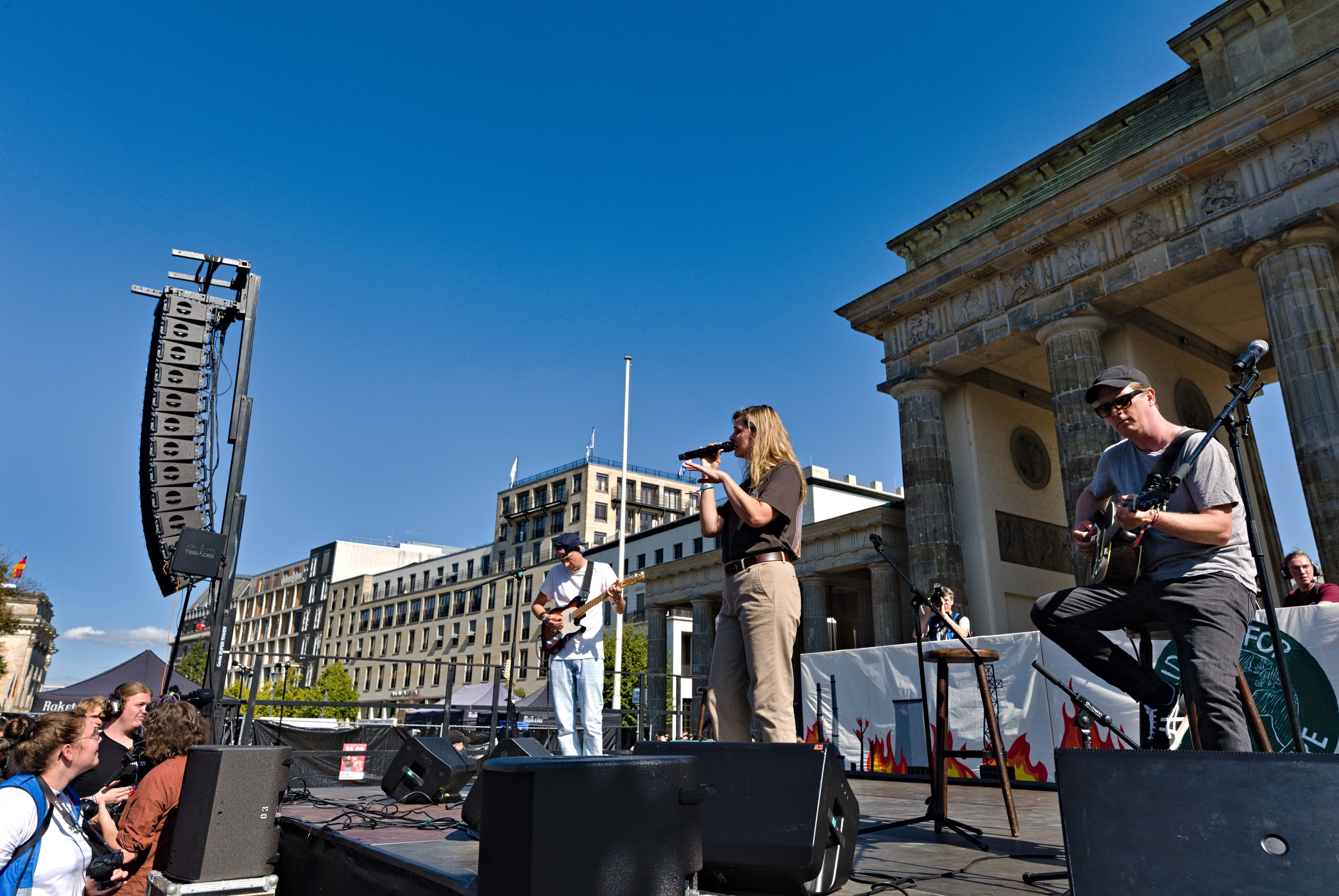 Eine Gruppe von Menschen, die auf einer Bühne vor dem Brandenburger Tor in Berlin Musik machen, mit Lautsprechern und Equipment drumherum, vor einem klaren blauen Himmel.