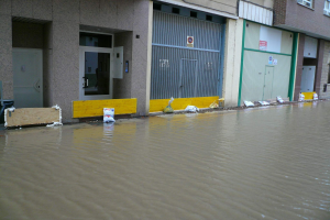Überschwemmte Straße mit Wasser, das ein Gebäude erreicht und die Umgebung sowie teilweise Fenster, Türen und andere Gegenstände bedeckt.
