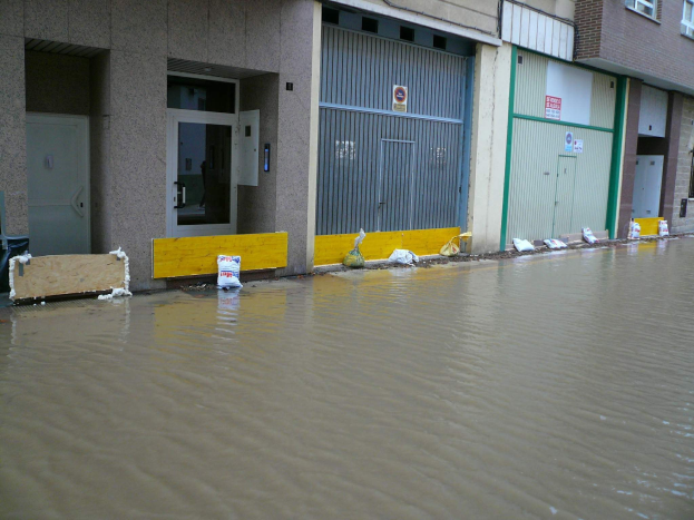Überschwemmte Straße mit Wasser, das ein Gebäude erreicht und die Umgebung sowie teilweise Fenster, Türen und andere Gegenstände bedeckt.