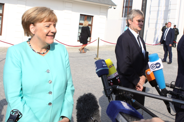 Bundeskanzlerin Angela Merkel beim Sprechen mit Reportern vor einem Gebäude, umgeben von Menschen mit Mikrofonen und einer Absperrung unter einem bewölkten Himmel.
