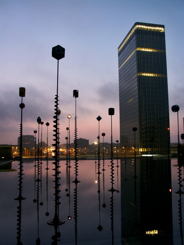 Eine nächtliche Stadtansicht mit mehreren Pfählen, Gebäuden und Lichtern, deren Spiegelungen auf einer nassen Oberfläche zu sehen sind, unter einem bewölkten Himmel.