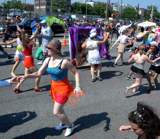 Eine Gruppe von Menschen, die eine Straße entlangmarschieren, einige mit Mötzen und Schirmen, andere tanzen, mit einem Banner, Verkehrskegeln, Fahrzeugen, Laternen, Schildern, Bäumen, Gebäuden und einem klaren blauen Himmel im Hintergrund.