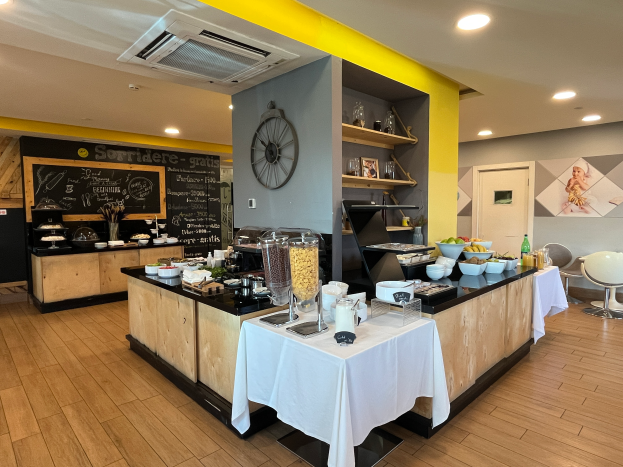 Ein Restaurant-Innenraum mit Tischen und Stühlen, Essensbehältern, einer Tafel mit Text, einer Wanduhr, Deckenleuchten, einer Klimaanlage und einer Tür im Hintergrund, befindet sich im Hôtel de Ville in Paris.