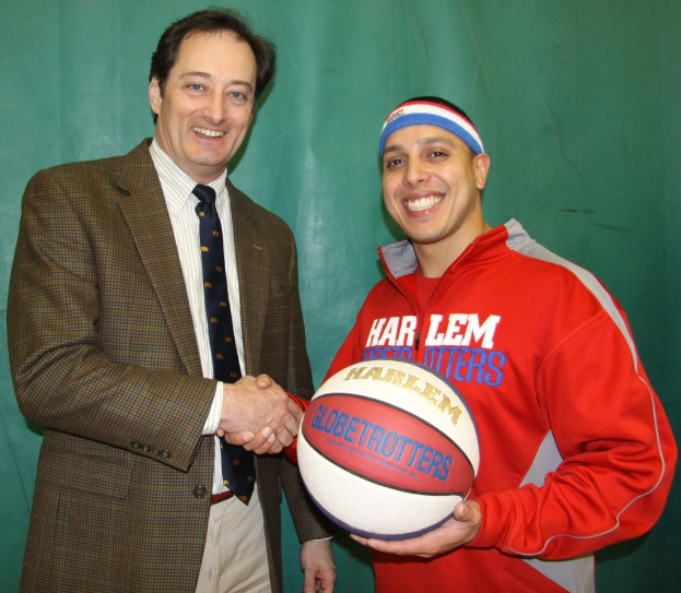 Zwei Männer, die sich die Hand geben, einer in einer roten Jacke mit einem Ball, der andere in einer grünen Blazer und blauer Krawatte, vor einem grünen Tuch stehend.