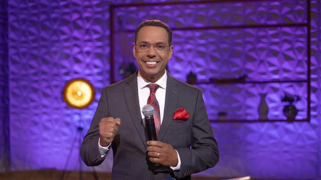 Stephen Colbert, Moderator von "The Daily Show with Trevor Noah", steht auf einer Bühne und hält ein Mikrofon, trägt einen Anzug, Krawatte und Brille, während im Hintergrund ein Scheinwerfer und ein Ständer zu sehen sind.