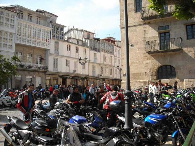 Ein cremefarbenes Gebäude mit zahlreichen Fenstern, das an ein Backsteingebäude mit einem Metallbalkon angrenzt, mit mehreren geparkten Fahrrädern und Menschen, die davor stehen und sich unterhalten.