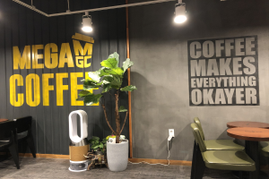 Ein Kaffeehaus-Interieur mit Tischen, Stühlen, einem "Mega Coffee"-Schild an der Wand, einem Topf mit einer Pflanze, einem Bodenreinigungsgerät und Deckenleuchten.