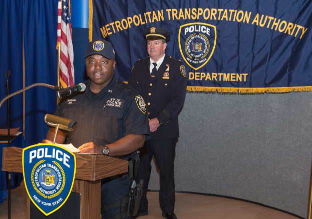 Zwei Polizeibeamte stehen vor einem Rednerpult, einer hölt Papiere und spricht in ein Mikrofon, mit einer Fahne, einem Banner und einem Tisch in der Nühhe, und einer Tafel mit Symbolen und Text im Hintergrund.