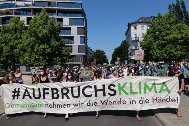 Gruppe von maskierten Menschen mit einem Banner 'Aufbruchsklima' vor einem Gebäude mit Bäumen und einem klaren Himmel.