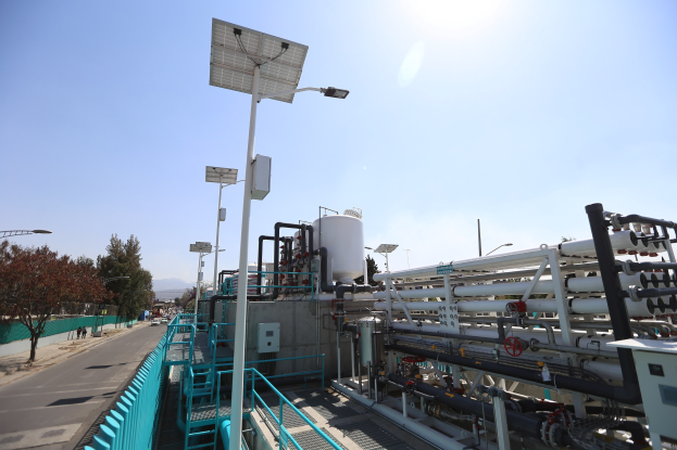 Großes industrielles Gebäude mit ausgedehnten Rohrleitungen und Solarpanelen auf dem Dach, umgeben von Sicherheitsgeländern, Versorgungsmasten, Straßenlaternen, Beschilderungen, Bäumen und Fahrzeugen auf einer nahen Straße, vor einem Hintergrund aus Hügeln und einem klaren blauen Himmel, Teil der neuen Wasseraufbereitungsanlage des California Department of Environmental Protection.