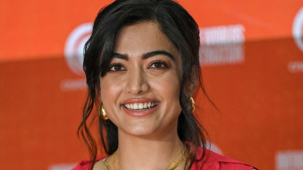 Eine Frau in einem leuchtend roten Kleid mit einem eleganten Dutt und dezentem Make-up posiert strahlend vor der Kamera, trägt schwarze High Heels und hat einen hellgelben Banner im Hintergrund mit der Aufschrift "Sonam Kapoors beste Schönheits Looks vom Cannes Film Festival."