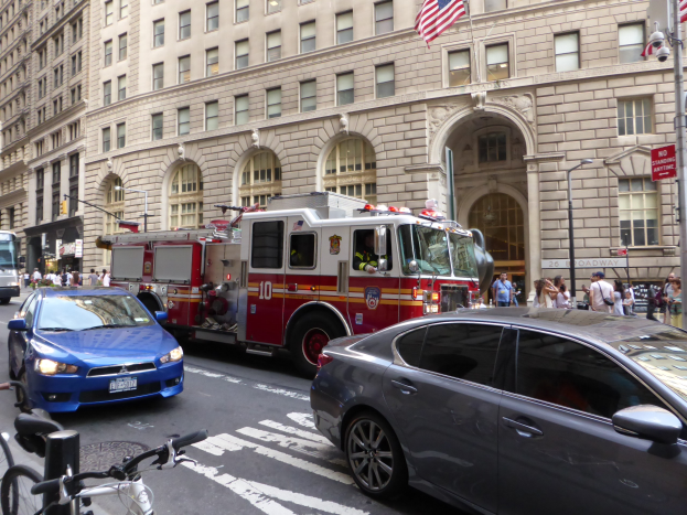 Ein rotes Feuerwehrauto auf einer Stra├če in New York City mit fahrenden Fahrzeugen, Passanten auf dem Gehweg, parkenden Fahrr├Ądern und Geb├Ąuden im Hintergrund.