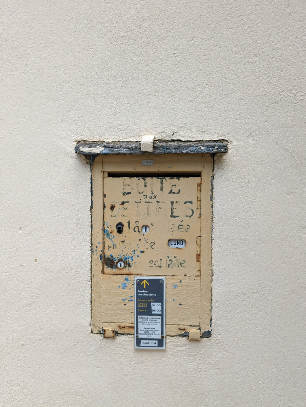 Alte rechteckige Metallklingel mit einem Griff oben, Schlüsselloch in der Mitte und einem Schild auf der Vorderseite, an einer verwitterten Gebäudewand mit sichtbaren Rissen und Absplitterungen montiert.