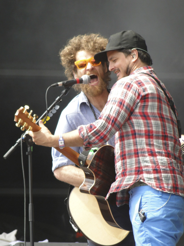 Zwei Männer auf der Bühne mit Gitarren und Mikrofonen, einer in rot-blau kariertem Hemd und blauen Hosen, der andere mit Goggles und schwarzer Mütze.
