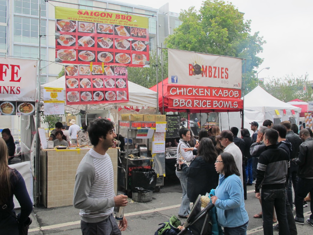 Menschen auf einer Straße vor der Stadt, mit einem Mann in einem grauen und weißen T-Shirt, der eine Wasserflasche hält, und einer Frau in einer blauen Jacke, die einen Kinderwagen schiebt, Stände mit weißen und roten Zelten, farbige Banner und im Hintergrund Gebäude, Bäume und eine Straßenlaterne.