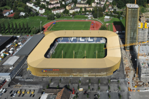 Luftaufnahme eines Fußballstadions im Zentrum einer Stadt, umgeben von Gebäuden, Bäumen, Straßen, Fahrzeugen, Pfählen und Kränen, mit saftigem Grün.