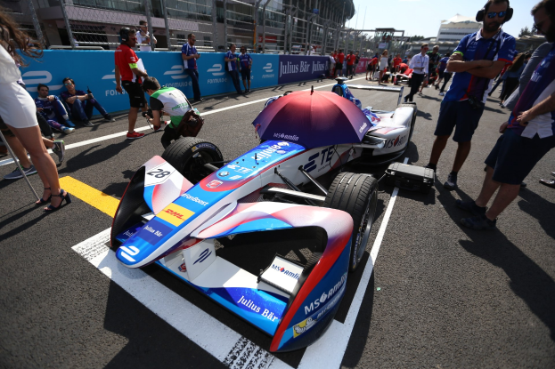 Formula-E-Auto mit weißer, blauer, roter und schwarzer Lackierung auf einer Rennstrecke geparkt, umgeben von Menschen, mit einem Schirm im Vordergrund und Rennstrecken-Infrastruktur im Hintergrund unter einem klaren blauen Himmel.