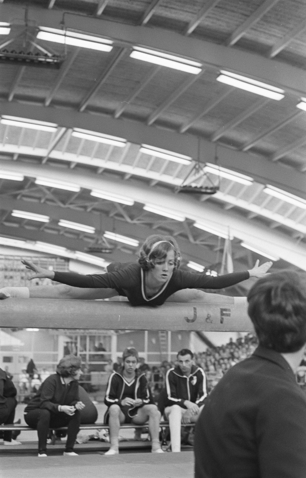 Eine Frau führt Gymnastik auf einem Balancebeam in einer Turnhalle durch, umgeben von Zuschauern auf Bänken und einigen Stehenden unter Deckenbeleuchtung, dargestellt in Schwarz-Weiß.