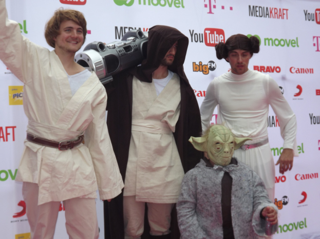 Eine Gruppe von Menschen in Star-Wars-Kostümen posiert auf einem roten Teppich, wobei einer ein Objekt und ein Banner mit Text und Logos im Hintergrund hält.